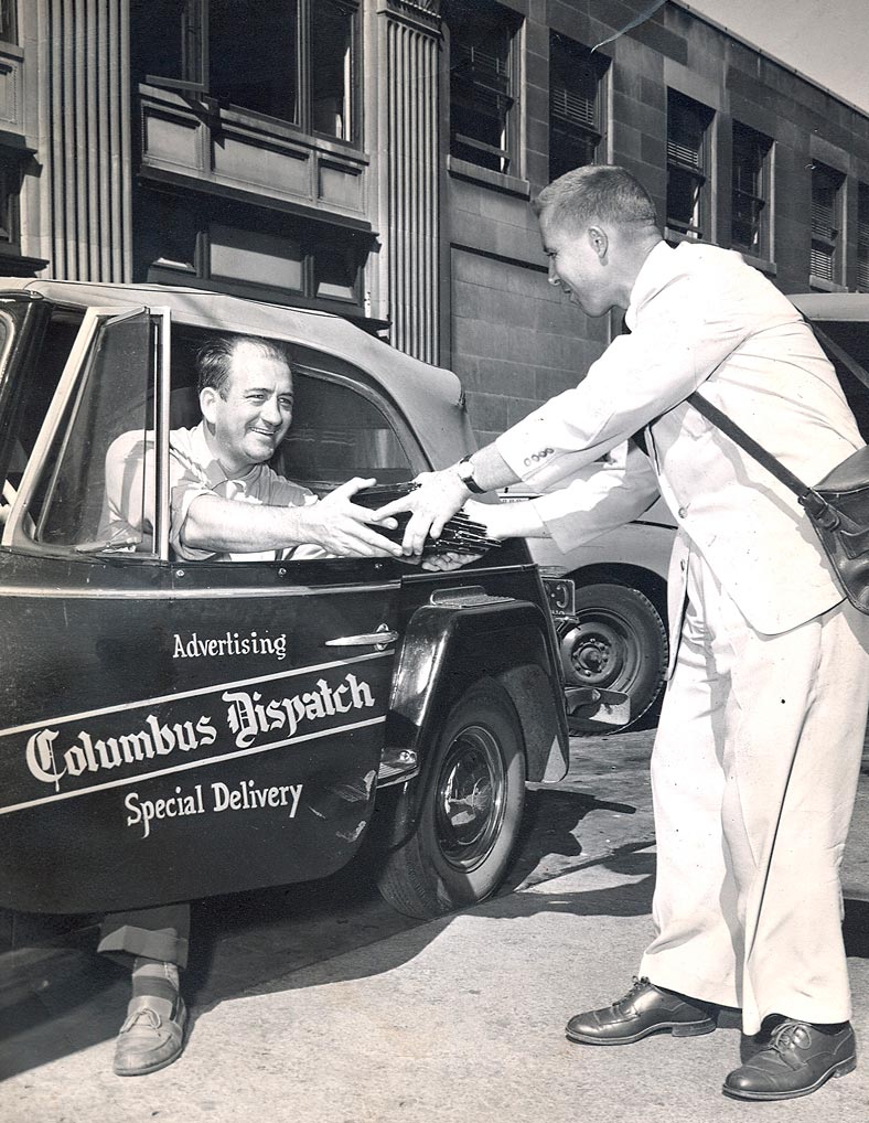 A man in a white suit hands an envelope to a smiling driver sitting in a car labeled “Columbus Dispatch Advertising Special Delivery” on a city street.