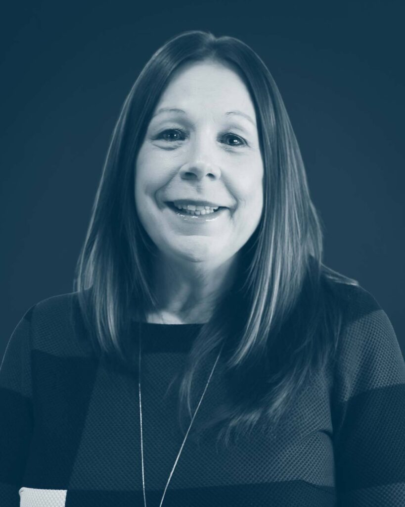 A woman with long, straight brown hair and a friendly smile, wearing a dark patterned top and a necklace, poses against a solid dark background. The image has a blue-toned filter.