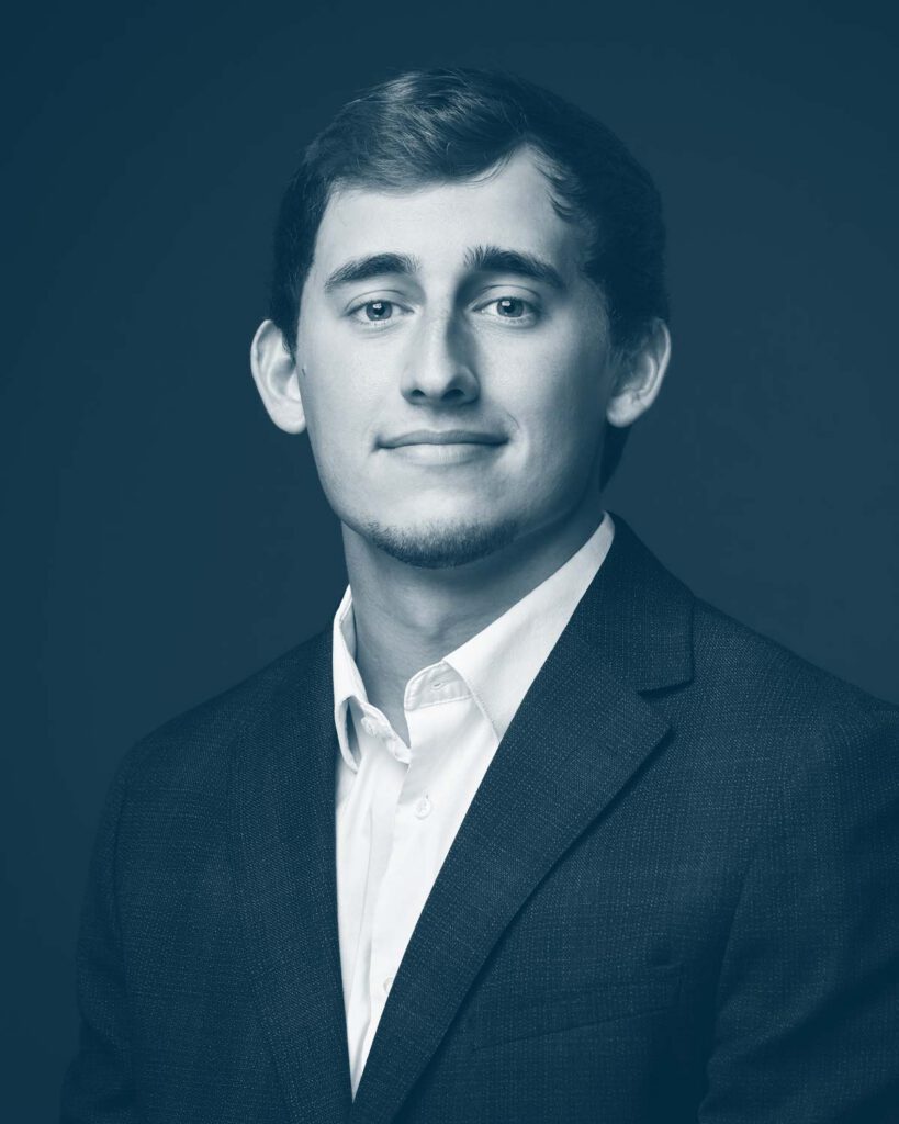 A young man with short hair wearing a dark suit jacket and a white shirt, posing for a professional portrait against a plain background. The image is in a blue monochrome tone.