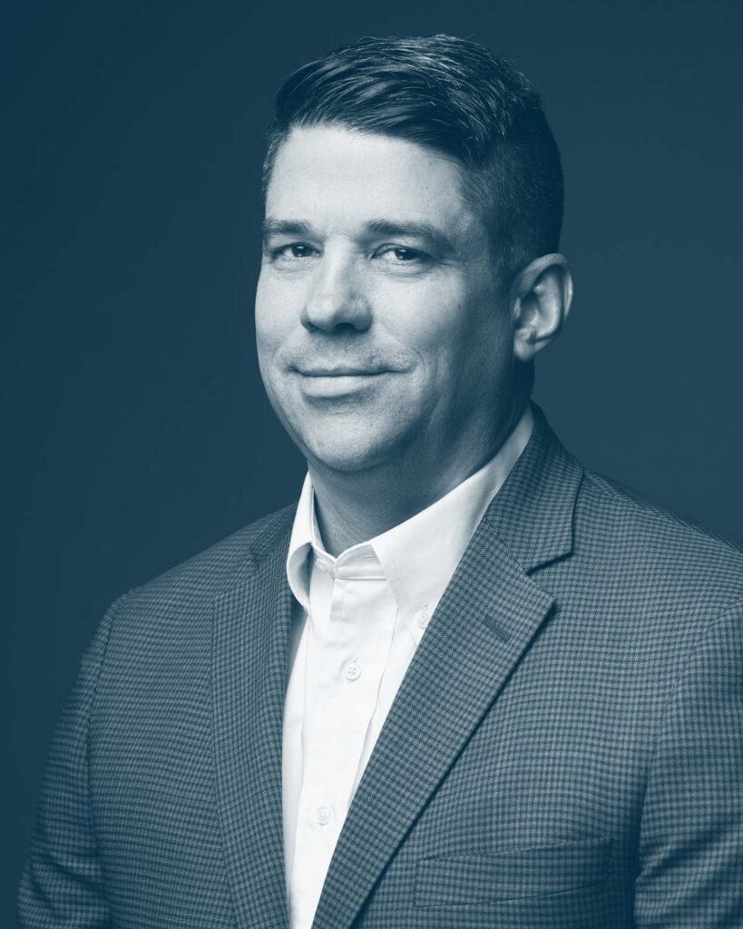 A man wearing a checkered suit jacket and white dress shirt smiles slightly while looking at the camera, against a plain background, in a blue-tinted portrait.