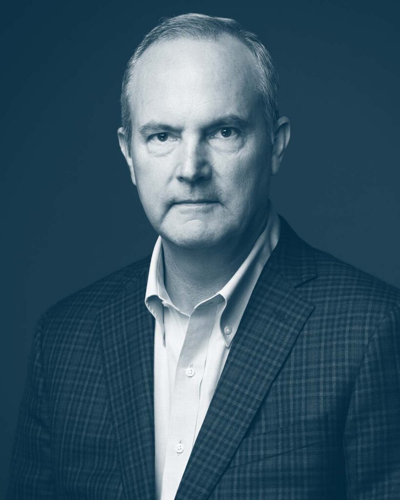 A middle-aged man with short hair wearing a plaid suit jacket and white dress shirt, looking seriously at the camera against a plain, dark background. The image has a blue monochrome filter.