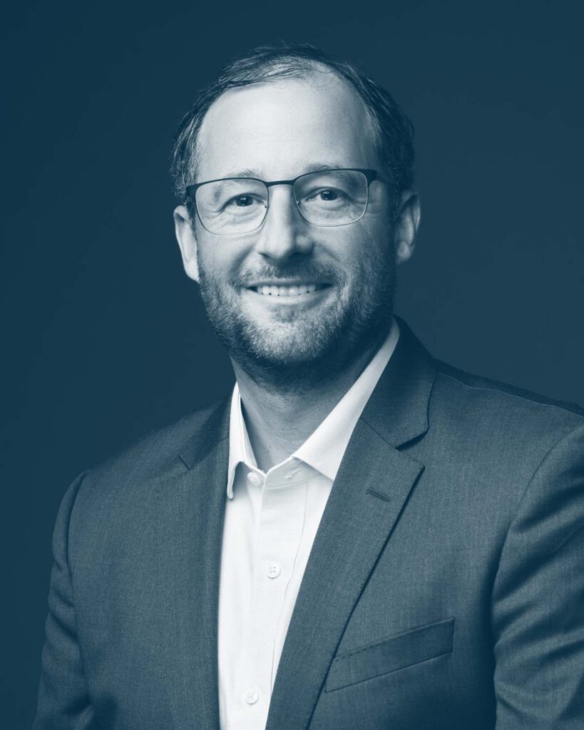 A man wearing glasses and a suit jacket over a white shirt is smiling at the camera against a dark background. The image is in blue monochrome.