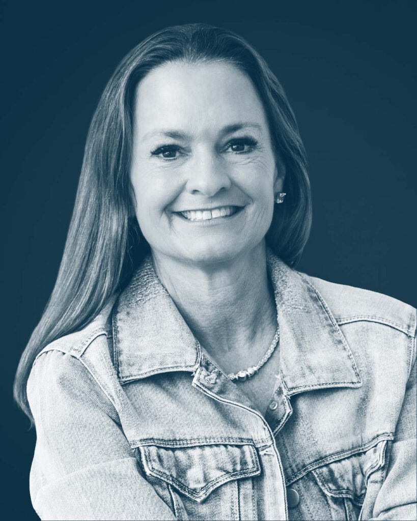 A woman with long straight hair smiles at the camera. She is wearing a denim jacket over a top and a necklace. The photo is in a blue monochrome style with a dark background.