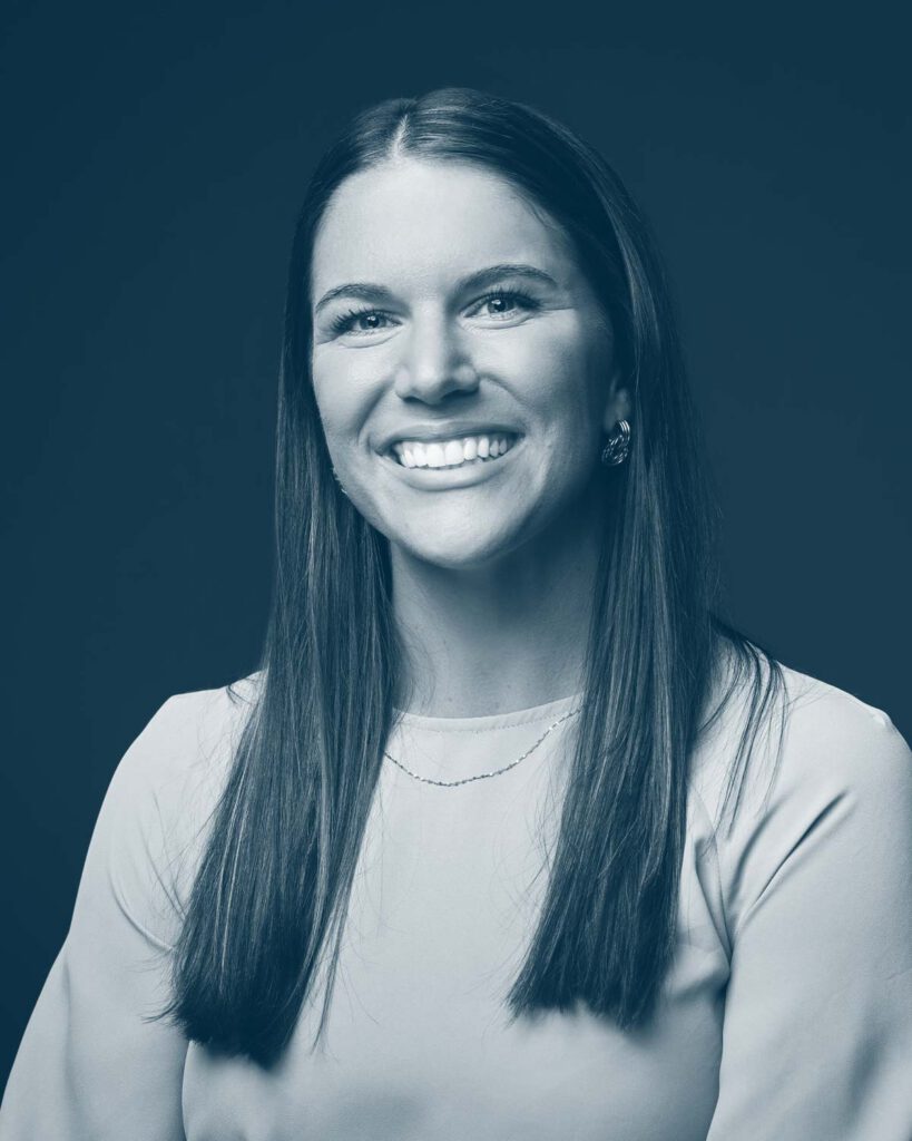 A woman with long straight hair, wearing a light-colored top and earrings, smiles while posing against a plain dark background in a studio portrait. The image is in blue monochrome.