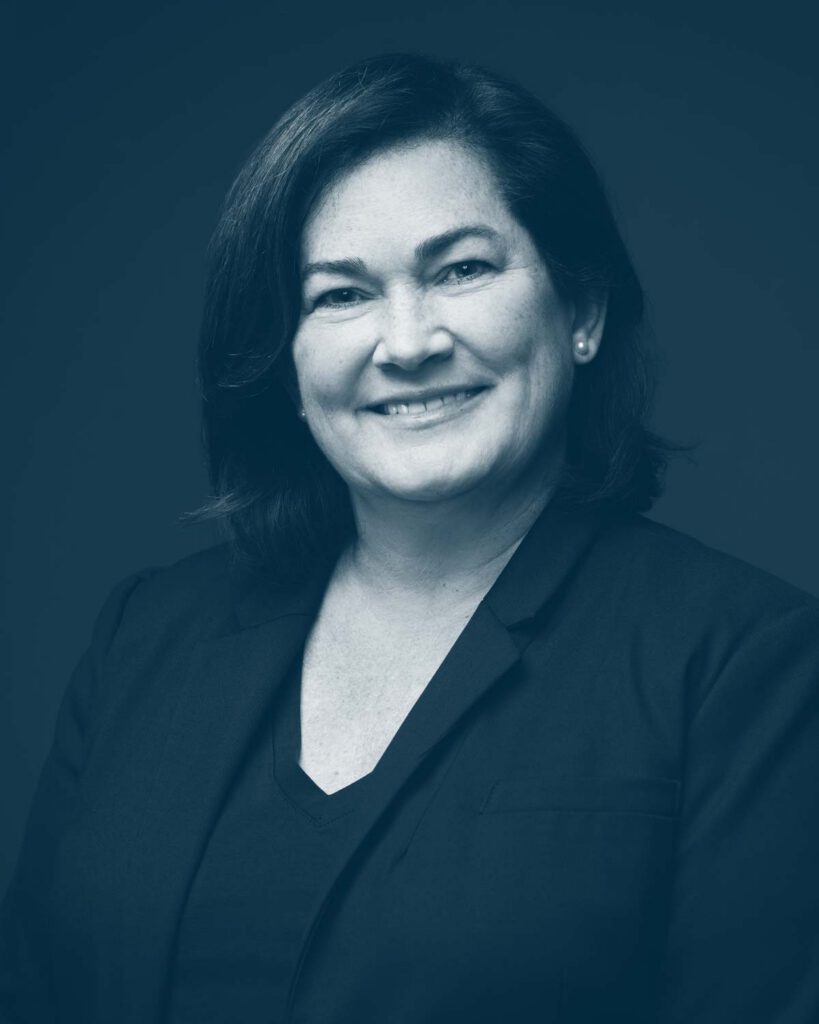 A woman with dark shoulder-length hair, wearing a dark blazer and a V-neck top, smiles at the camera against a plain, dark background. The image is in a blue monochrome tone.