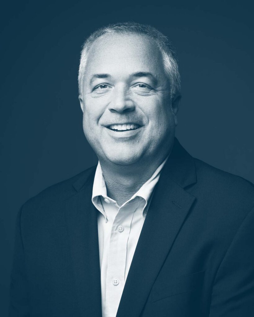 A smiling man with short gray hair wearing a suit jacket and collared shirt, posing in front of a plain background. The image is in a blue monochrome tone.