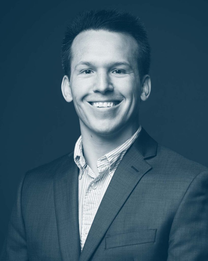 A young man wearing a suit jacket and striped shirt smiles at the camera in this professional studio portrait with a blue-toned filter.