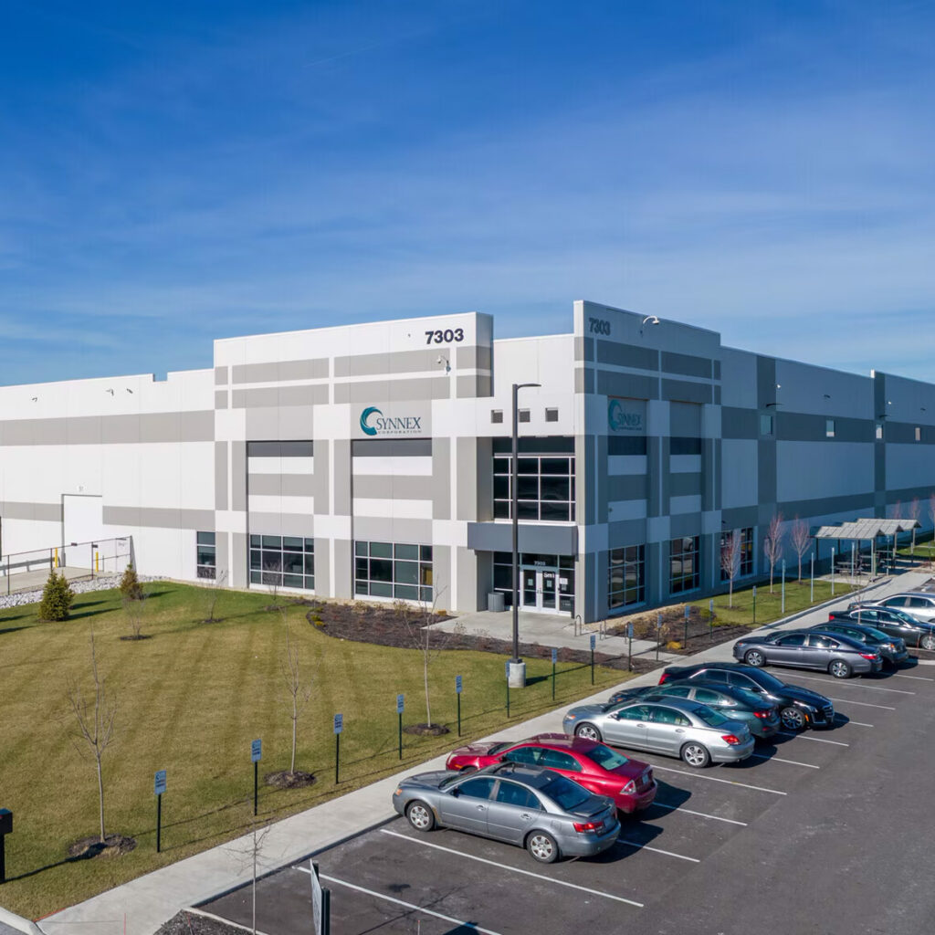 A large, modern industrial building with “Annex” signage and address 7303, surrounded by a parking lot with several parked cars and a grassy area, under a clear blue sky.