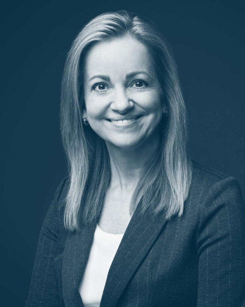 A woman with long, straight hair, wearing a pinstripe blazer and a light top, smiles at the camera against a dark background. The image is in a blue monochrome tone.
