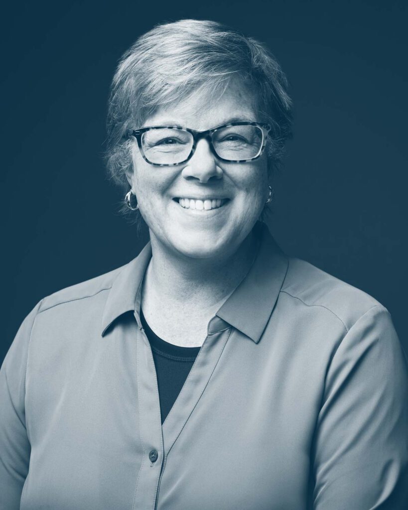 A middle-aged woman with short, light hair and glasses smiles at the camera. She is wearing a button-up shirt over a dark top and hoop earrings. The image has a blue-tinted monochrome effect.