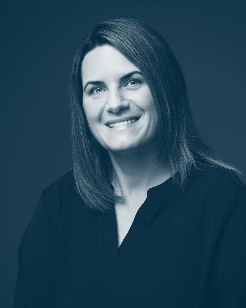A woman with straight shoulder-length hair, wearing a dark top, smiles at the camera against a plain background. The image has a blue monochrome filter.
