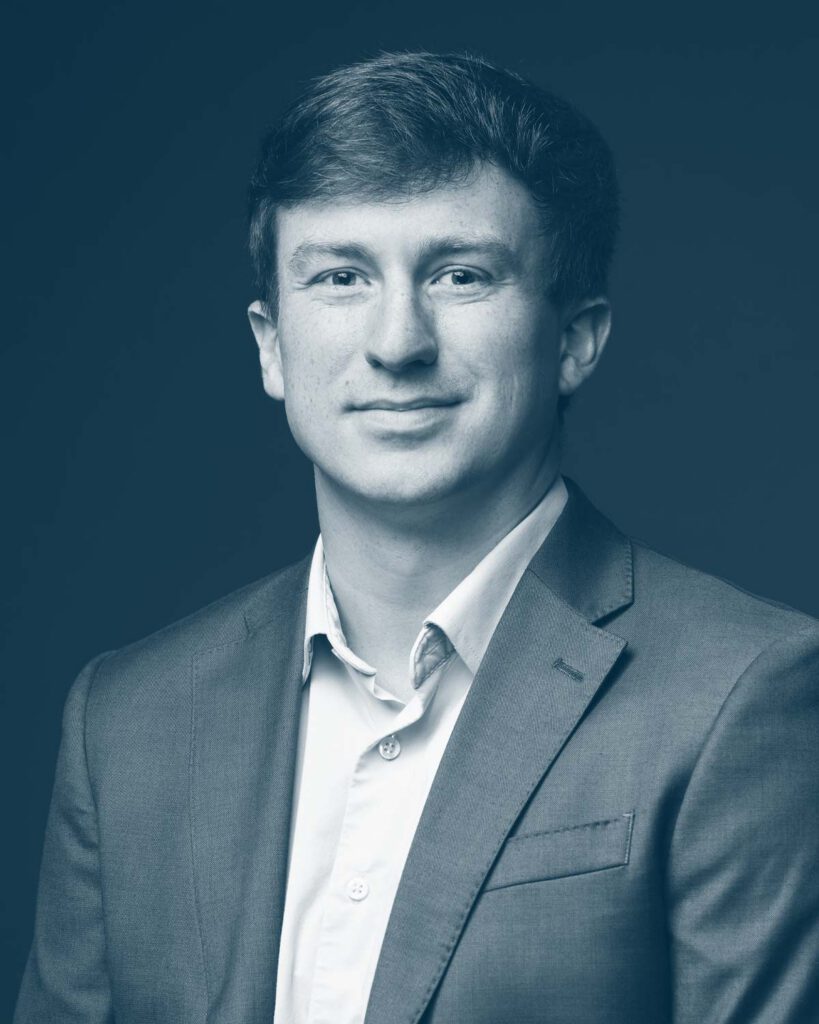 A young man with short hair wearing a suit jacket and dress shirt, looking at the camera with a slight smile against a plain dark background. The image is in monochrome blue tones.