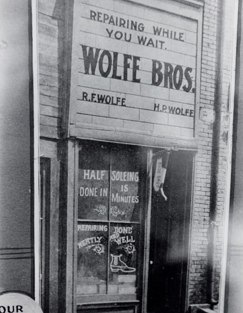 Black and white photo of a storefront with a sign reading “WOLFE BROS. REPAIRING WHILE YOU WAIT.” Windows advertise shoe repair services like “HALF SOLEING DONE IN 15 MINUTES” and “REPAIRING NEATLY DONE WELL.”.