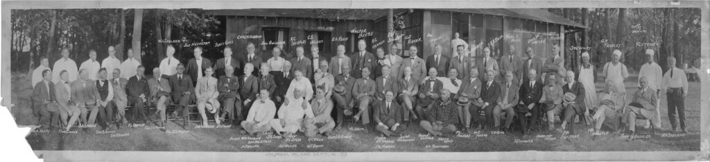 Black and white panoramic photo of a large group of men posing outdoors in front of a building, with names handwritten above their heads. Most are in suits or white clothing, some are seated, others stand behind. Trees are visible in the background.