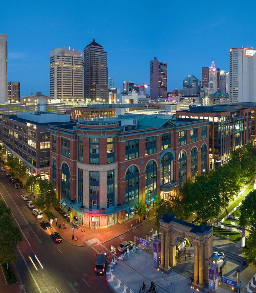 Aerial view of downtown Columbus, Ohio at dusk, featuring tall buildings, a large brick building with arched windows, busy streets, and a park with a lit-up archway entrance.