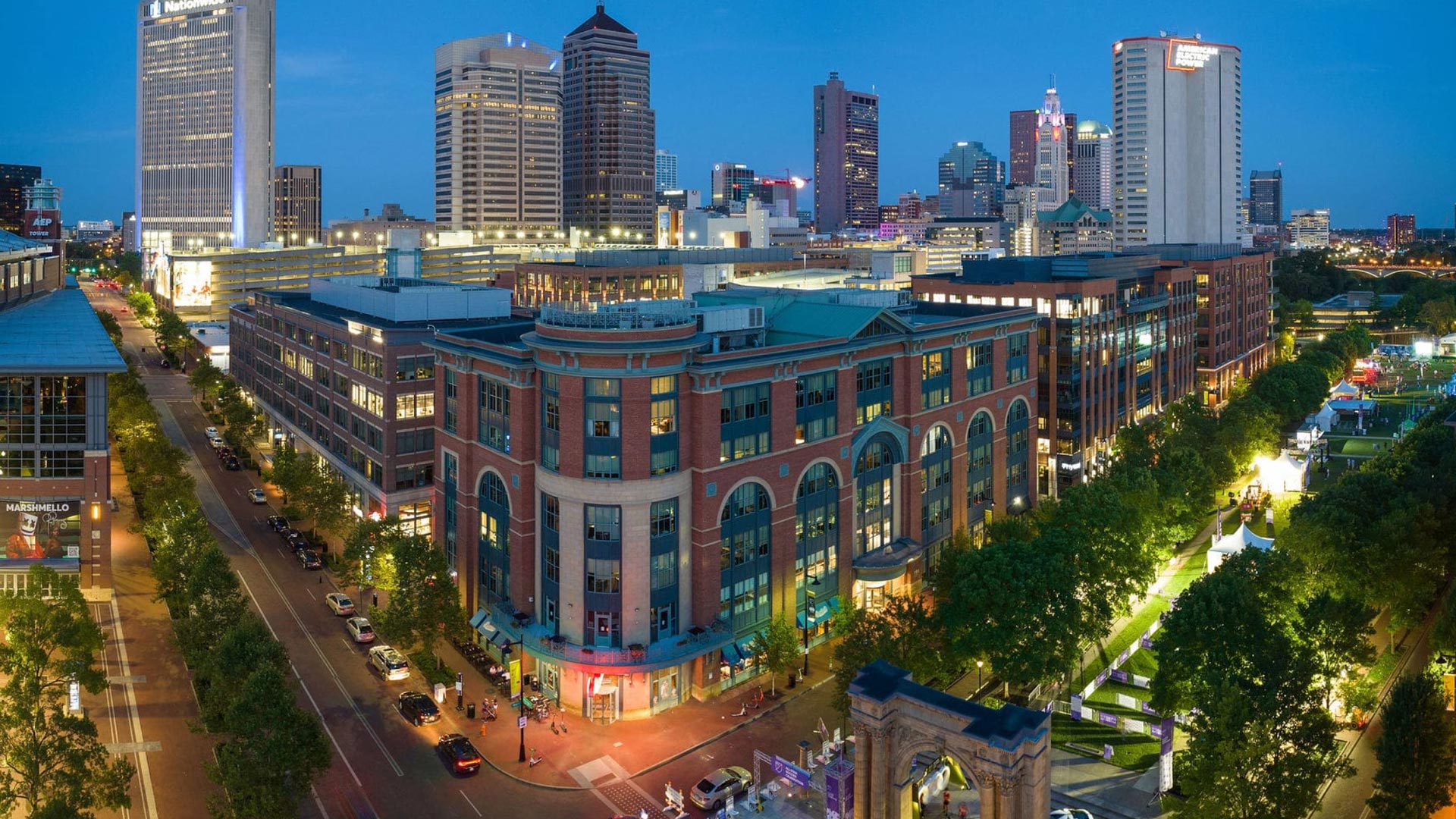 Aerial view of a lively downtown cityscape at dusk, featuring modern and historic buildings, busy streets with cars, and greenery along the sidewalks, all illuminated by streetlights and city lights.