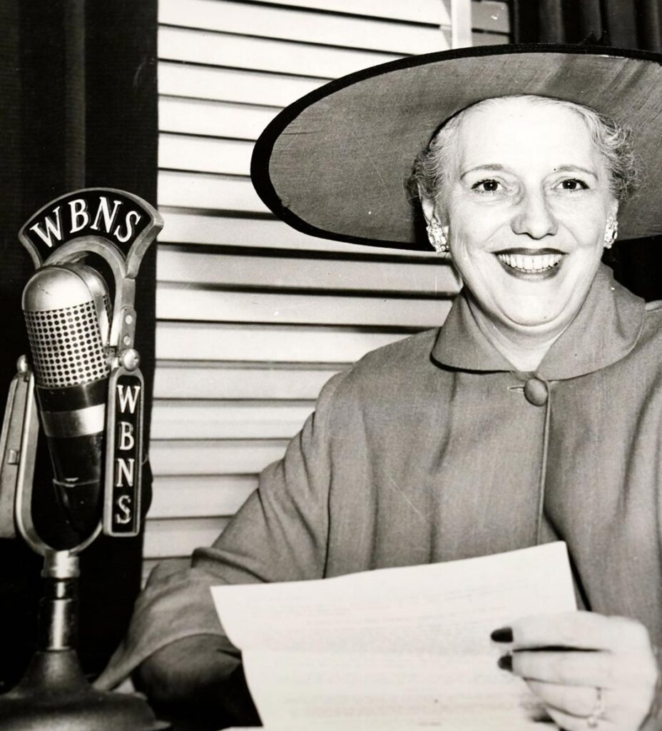 A woman in a wide-brimmed hat and coat smiles while sitting at a desk with papers, speaking into a vintage WBNS microphone. The background shows horizontal blinds. The photo is black and white.