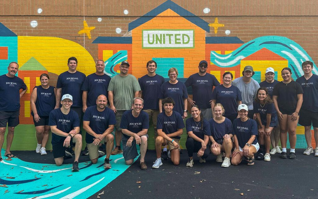 A group of people pose and smile in front of a colorful mural with the word UNITED on it. Most are wearing matching dark blue JOURNAL shirts, and the group is standing and kneeling outdoors on pavement.