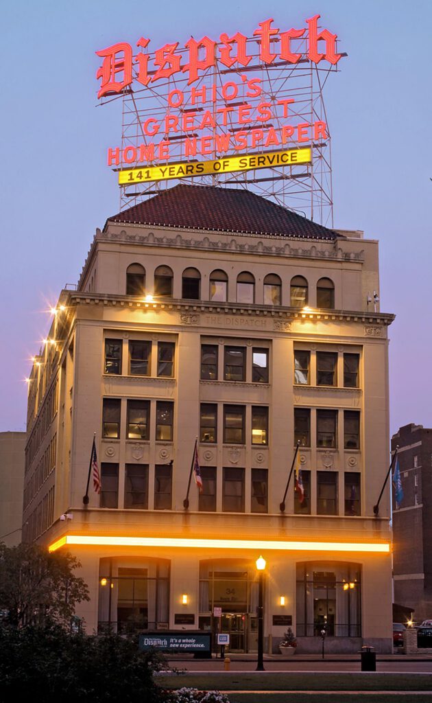 Historic building with a large lit-up sign on the roof reading Dispatch Ohios Greatest Home Newspaper, 141 Years of Service. The building is beige with large windows and flags at the entrance.