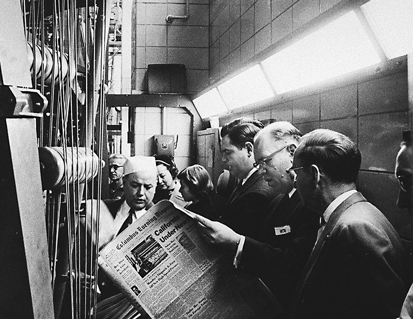 A group of men in suits examine machinery in an industrial setting while one man reads a newspaper with the headline “Cathie Under Fire.” Bright overhead lights illuminate the tiled room.