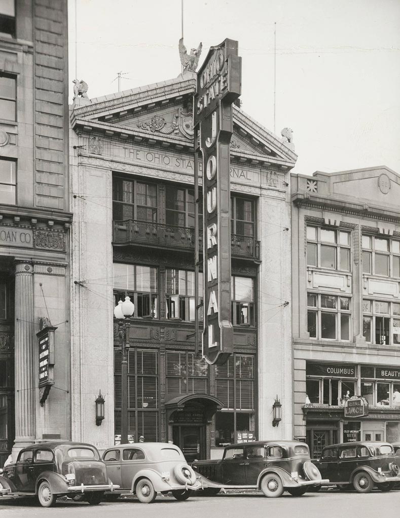 Black-and-white photo of a historic city street with parked vintage cars and the Ohio State Journal building, featuring a large vertical sign that reads JOURNAL. Nearby are various storefronts and businesses.