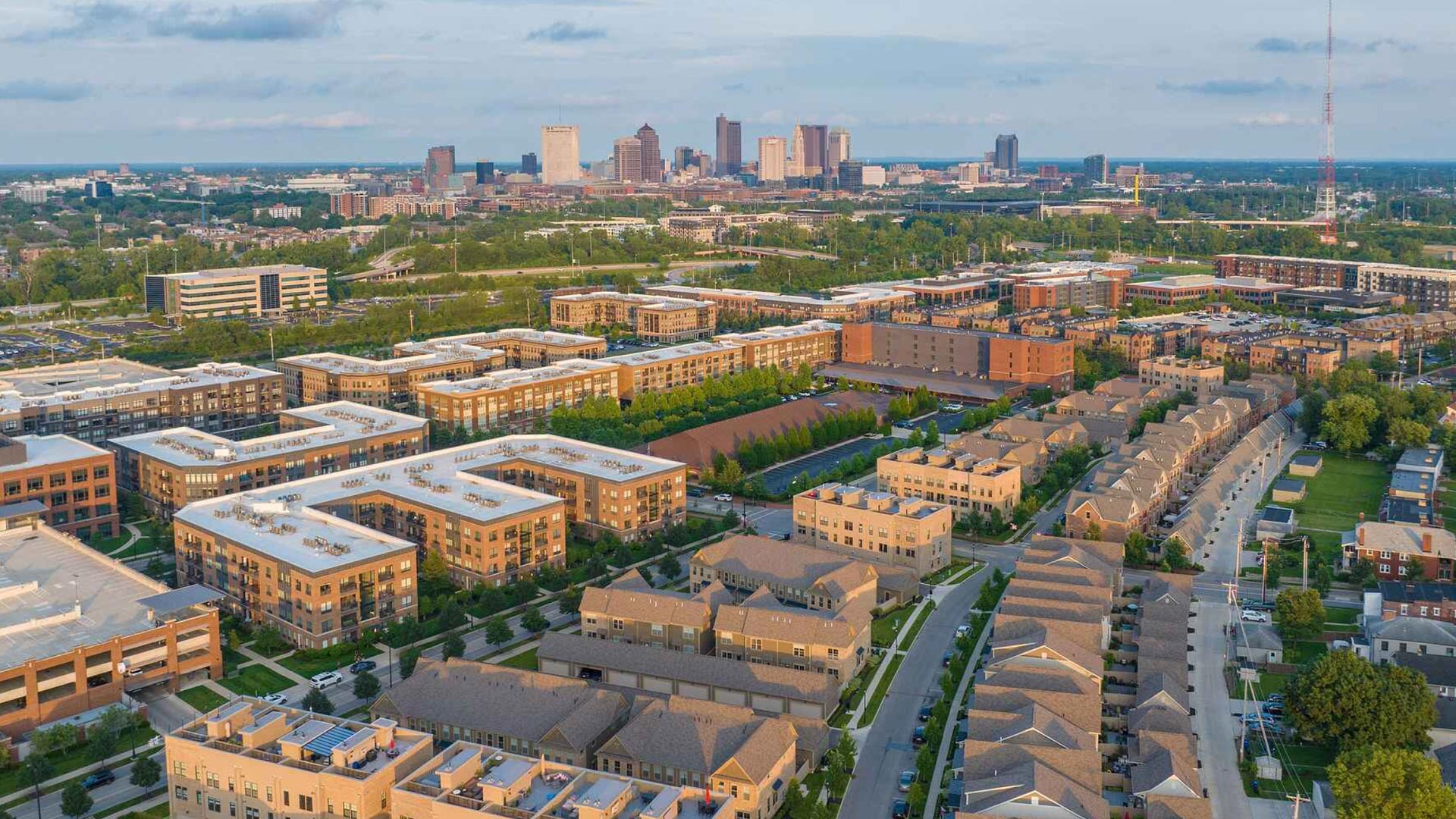 Aerial view of a suburban neighborhood with rows of houses and apartment buildings, trees, and streets, with a city skyline in the background under a partly cloudy sky.