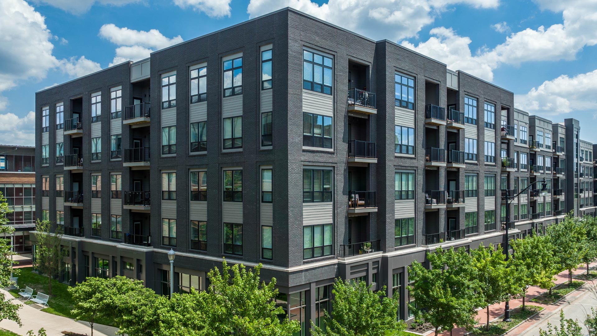 Modern four-story apartment building with dark gray brick exterior, large windows, and small balconies, surrounded by green trees and a clear blue sky with scattered clouds.