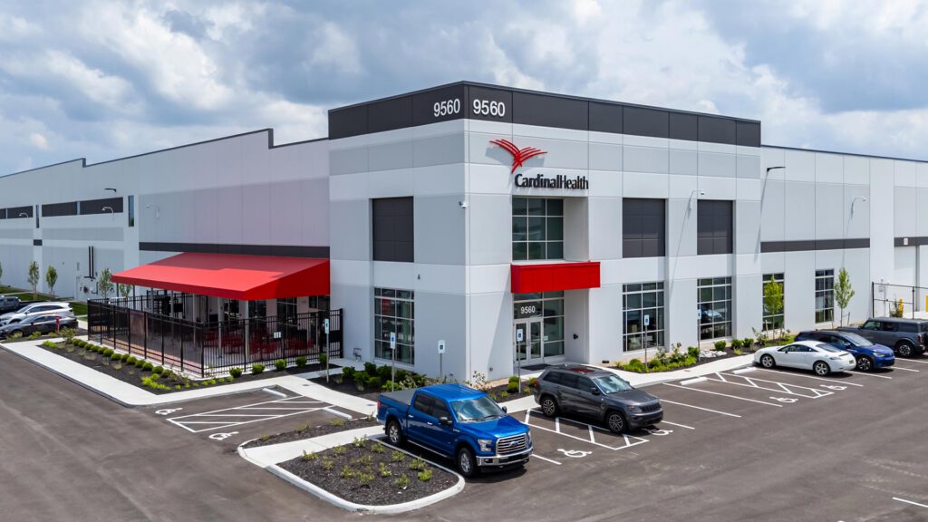 A modern Cardinal Health warehouse building with a red awning, multiple entrances, and parked vehicles in front under a partly cloudy sky. The address 9560 and Cardinal Health logo are visible on the building.