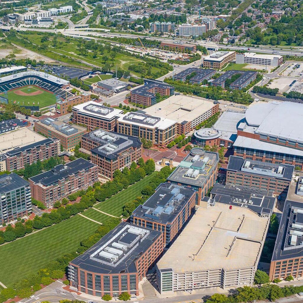 Aerial view of an urban area featuring multiple large office buildings, parking structures, green spaces, a baseball field, and surrounding roads with residential neighborhoods in the background.