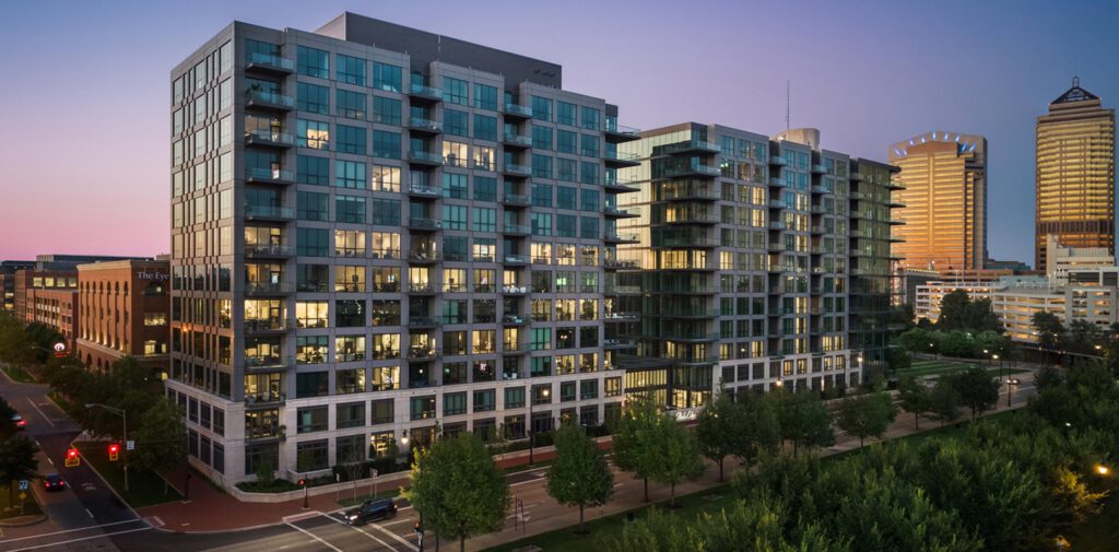 A modern, multi-story apartment building with glass balconies and illuminated windows stands in an urban area at sunset, surrounded by trees and nearby high-rise office buildings.
