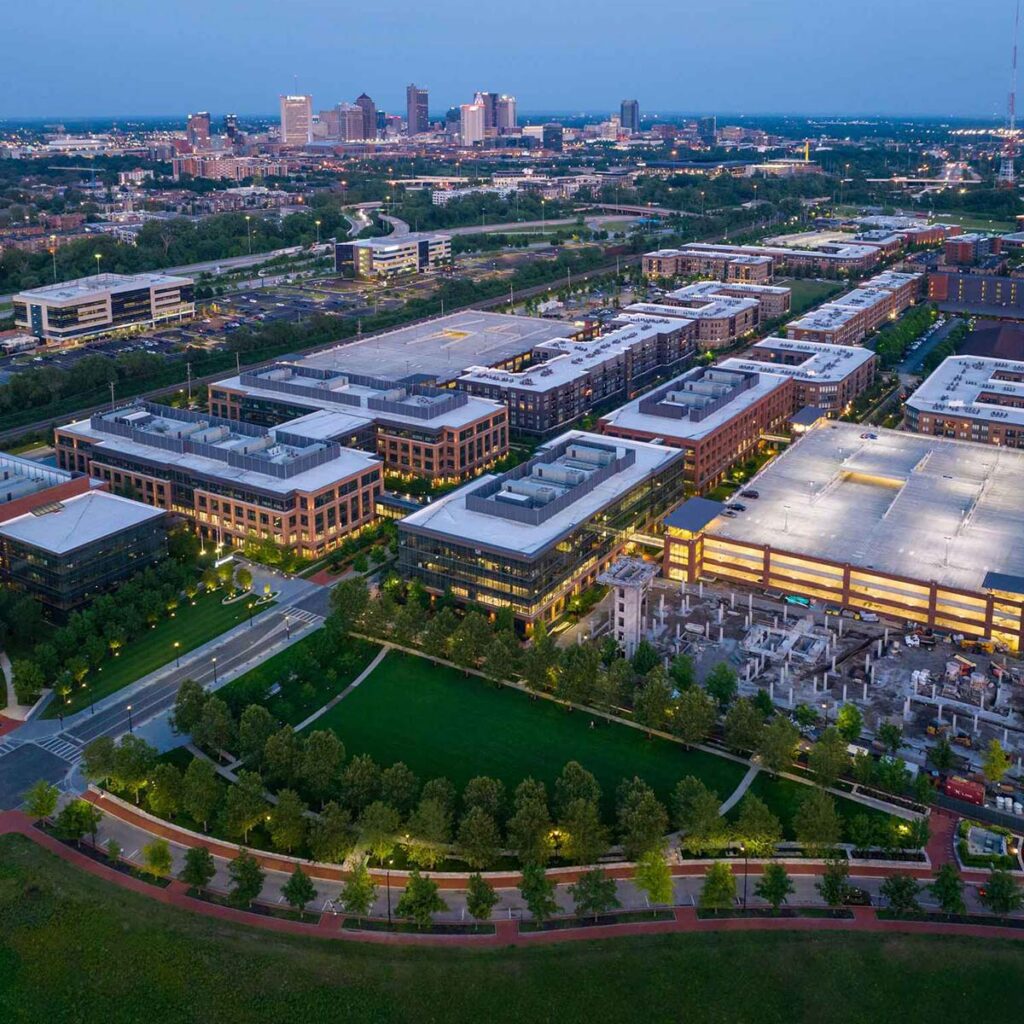 Aerial view of a modern urban district with office buildings, parking structures, green spaces, and tree-lined streets at dusk, with a city skyline visible in the background.
