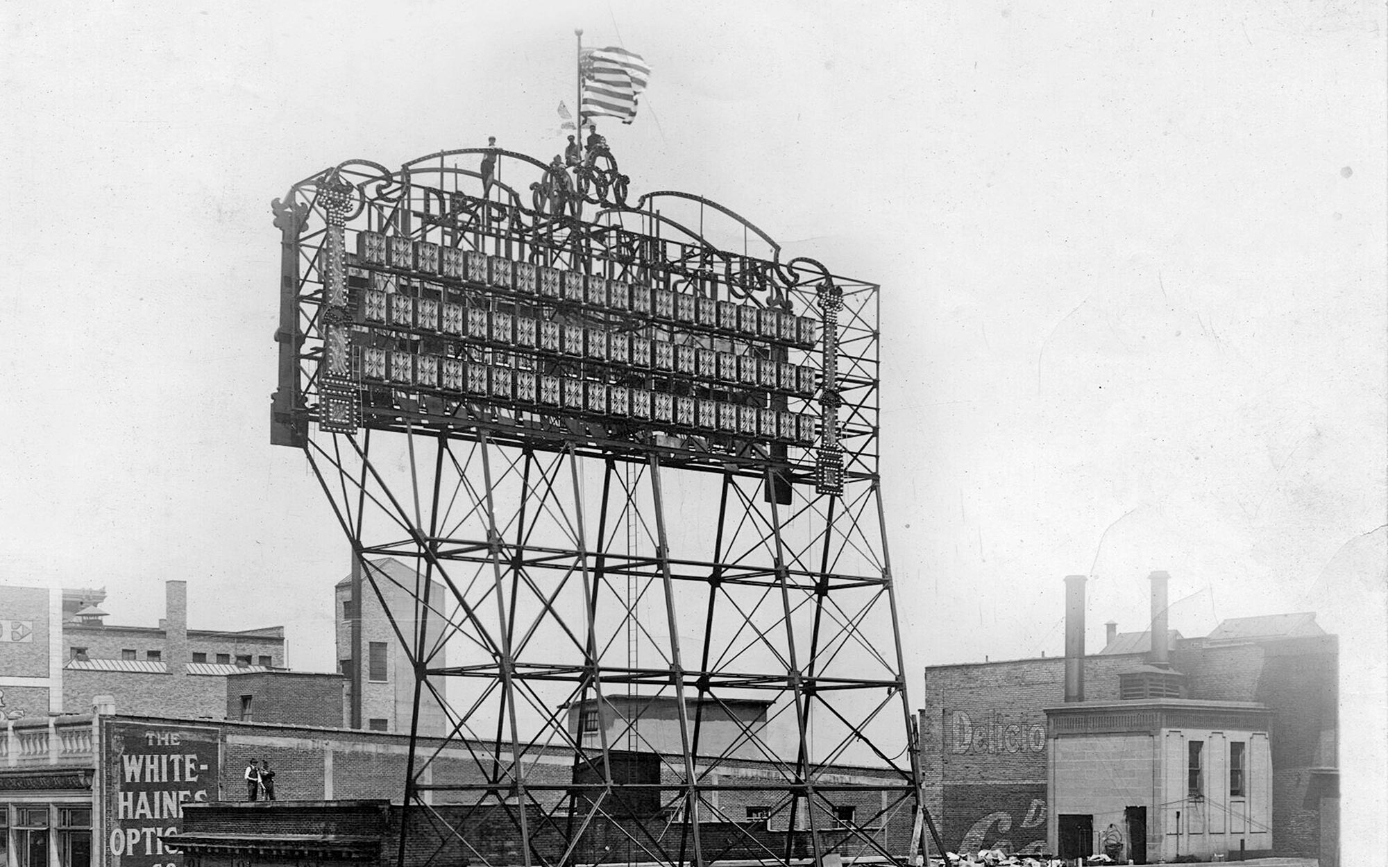 A large, vintage baseball scoreboard stands atop a tall metal frame above rooftops in a city. An American flag waves at the top, and the scoreboard displays game information in columns.