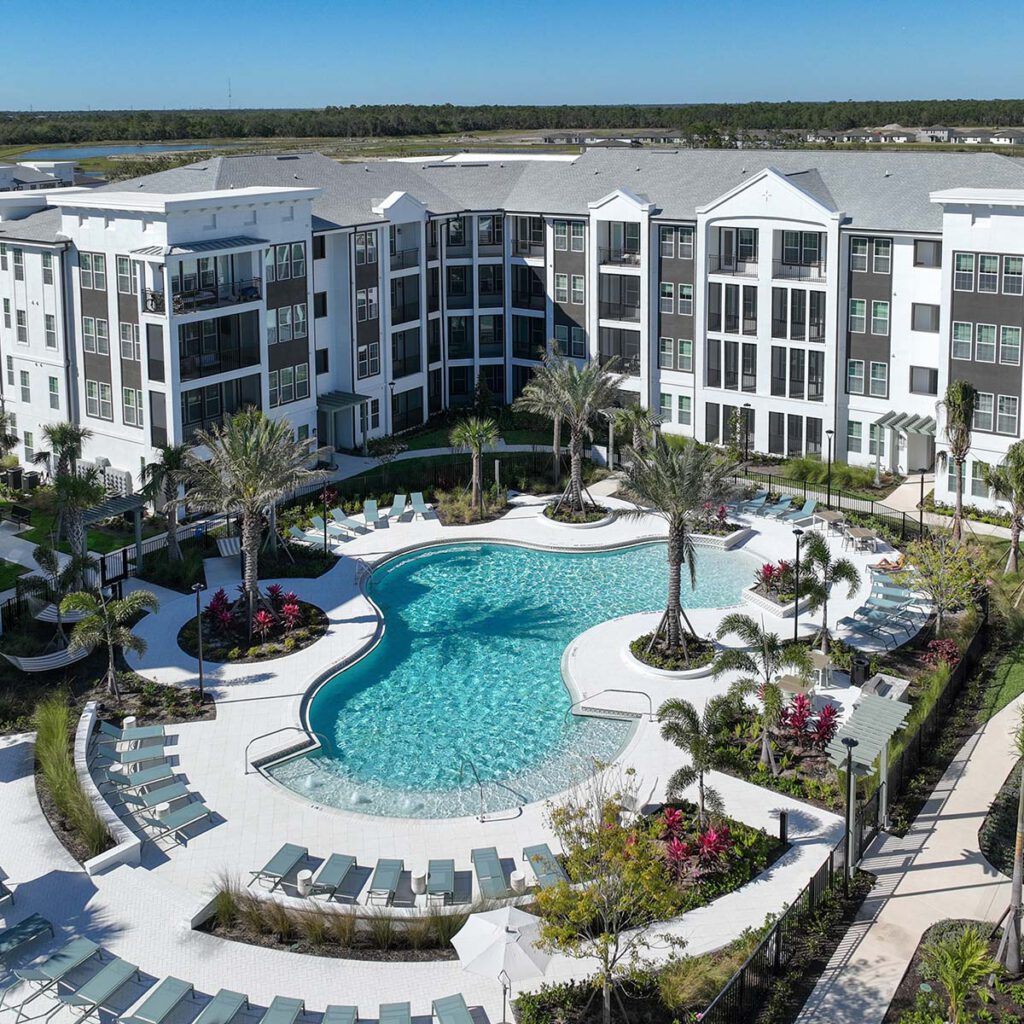 A large, modern apartment complex with white buildings surrounding a resort-style swimming pool, palm trees, lounge chairs, and landscaped gardens under a clear blue sky.