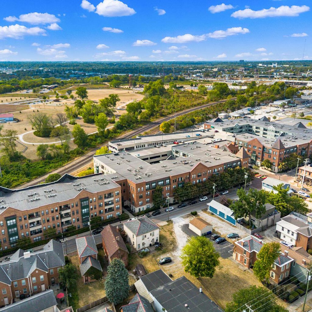 Aerial view of a suburban neighborhood with multi-story apartment buildings, smaller houses, green spaces, and a train track running alongside the area under a bright blue sky with scattered clouds.