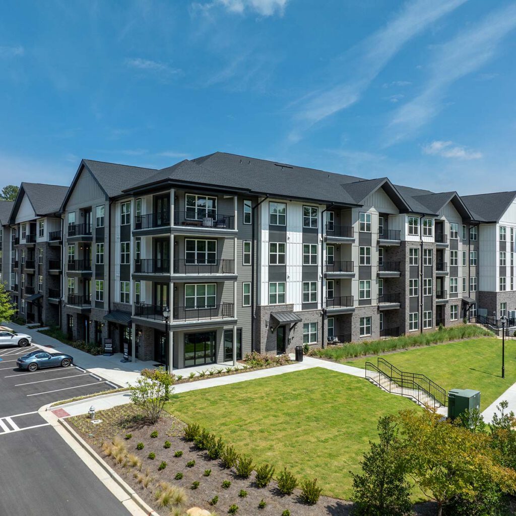A modern, three-story apartment building with balconies, large windows, and white and gray exterior, surrounded by green lawns, landscaping, and parked cars on a sunny day.