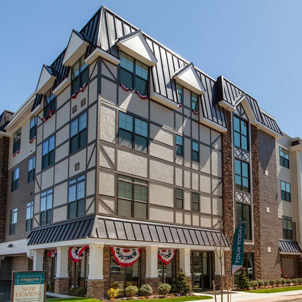A modern four-story apartment building with Tudor-style details, large windows, dark roof, and patriotic bunting on a sunny day. Signs read Now Leasing and Tour Today. Small shrubs and flowers line the entrance.