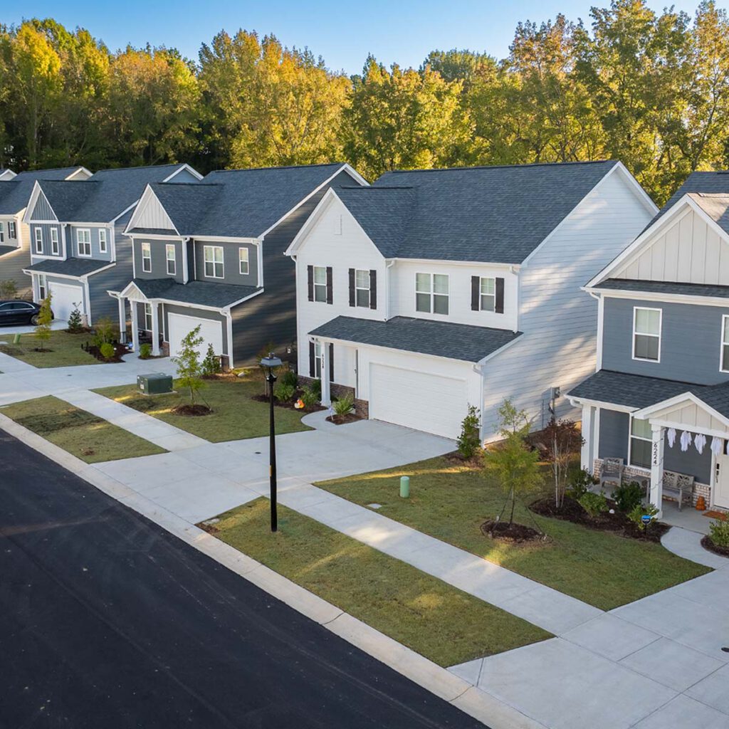 A row of modern, two-story suburban houses with gray siding, white trim, and neatly landscaped yards lines a clean, quiet street under a clear blue sky. Trees with green foliage are visible in the background.