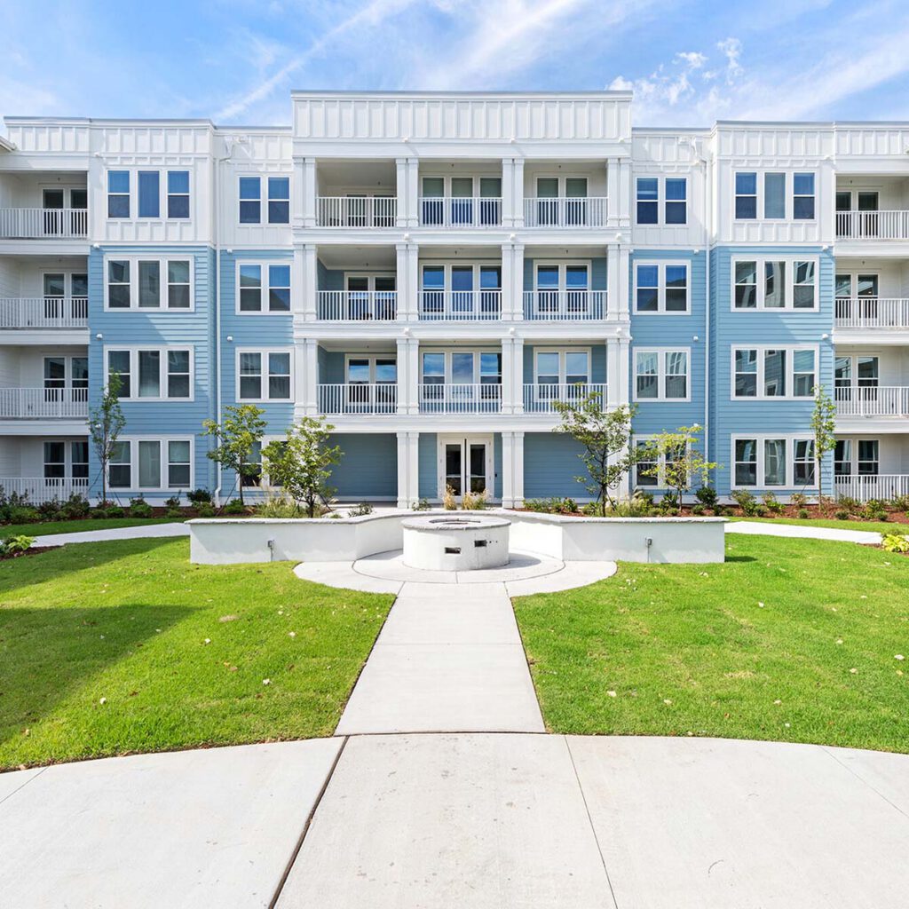 A modern, four-story apartment building with blue and white siding, multiple balconies, and large windows, surrounded by well-maintained green grass, landscaping, and a circular walkway with a central fountain.