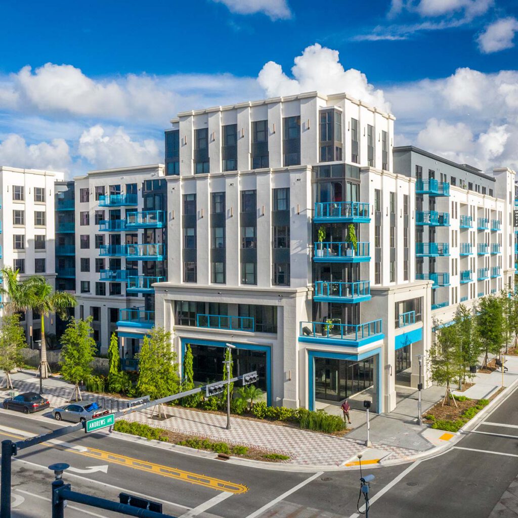 Modern mid-rise apartment building with white and gray exterior, blue balconies, and awnings. Trees line the street corner, with crosswalks and a street sign visible under a partly cloudy sky.