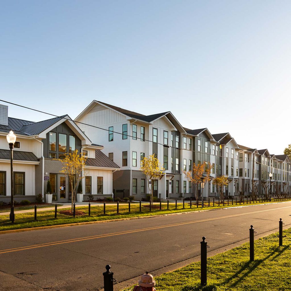 A modern, three-story apartment building with large windows and gabled roofs sits alongside a quiet street lined with young trees and a sidewalk on a sunny day.