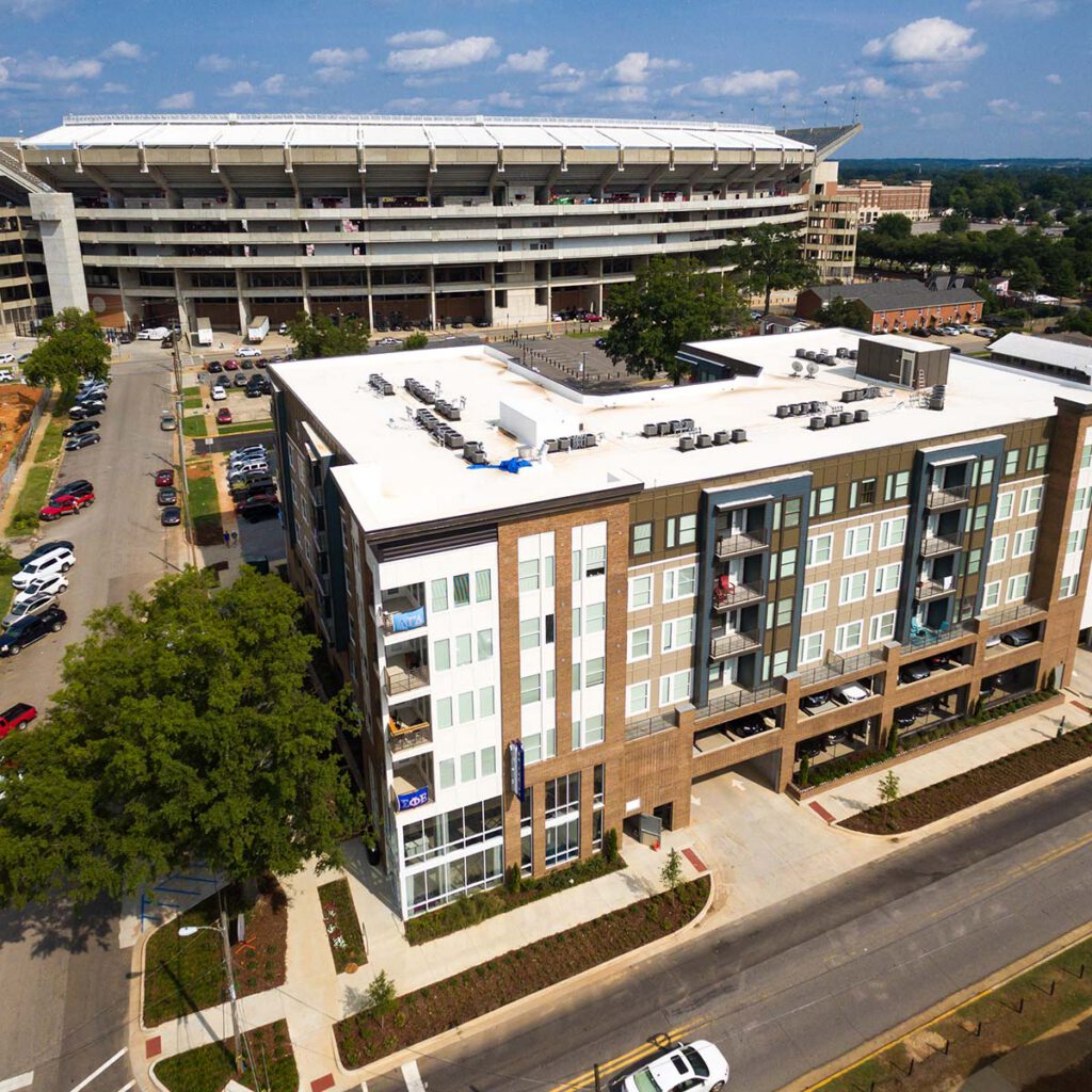 A multi-story apartment building with balconies is in the foreground, with a large football stadium and several parked cars visible in the background under a partly cloudy sky.