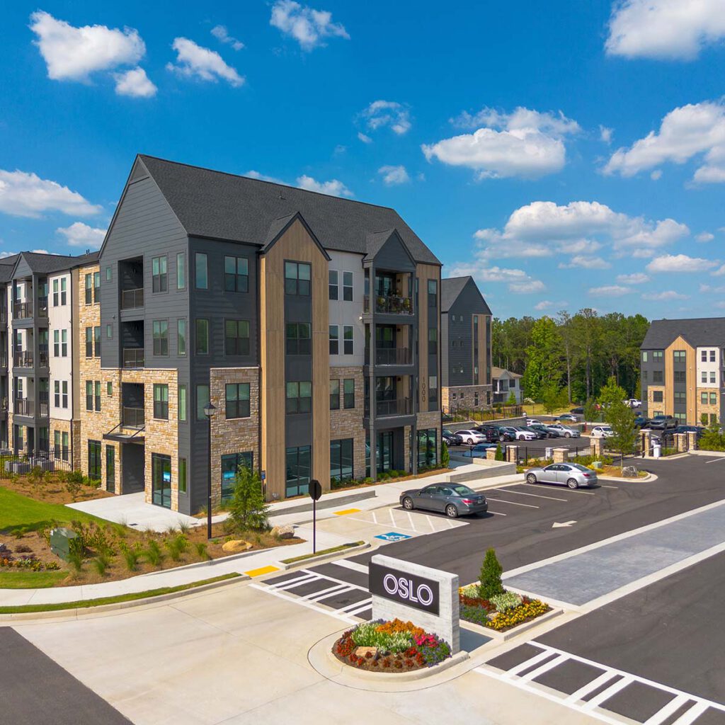 Modern apartment complex with dark roofs and stone accents, surrounded by landscaped greenery and parked cars. A sign reading OSLO is at the entrance under a blue sky with scattered clouds.