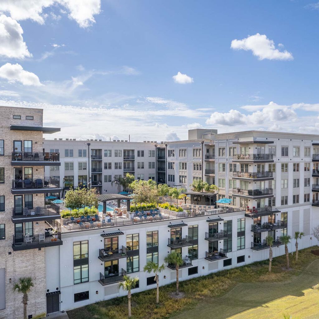 Modern apartment complex with multiple stories, balconies, and a rooftop pool area surrounded by trees and lounge chairs. The sky is bright with scattered clouds, and palm trees line the lawn below.