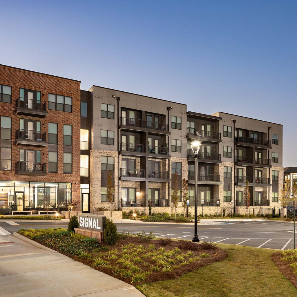Modern apartment complex with brick and light-colored façades, balconies, large windows, and landscaped grounds. A sign reading “SIGNAL” is near the entrance, and a streetlamp is lit at dusk.