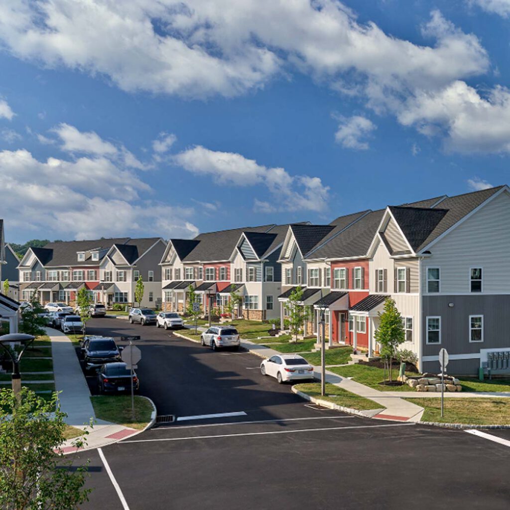 A residential neighborhood with modern two-story townhouses, neatly lined along a clean, curved street with parked cars, green lawns, and a bright blue sky with scattered clouds.