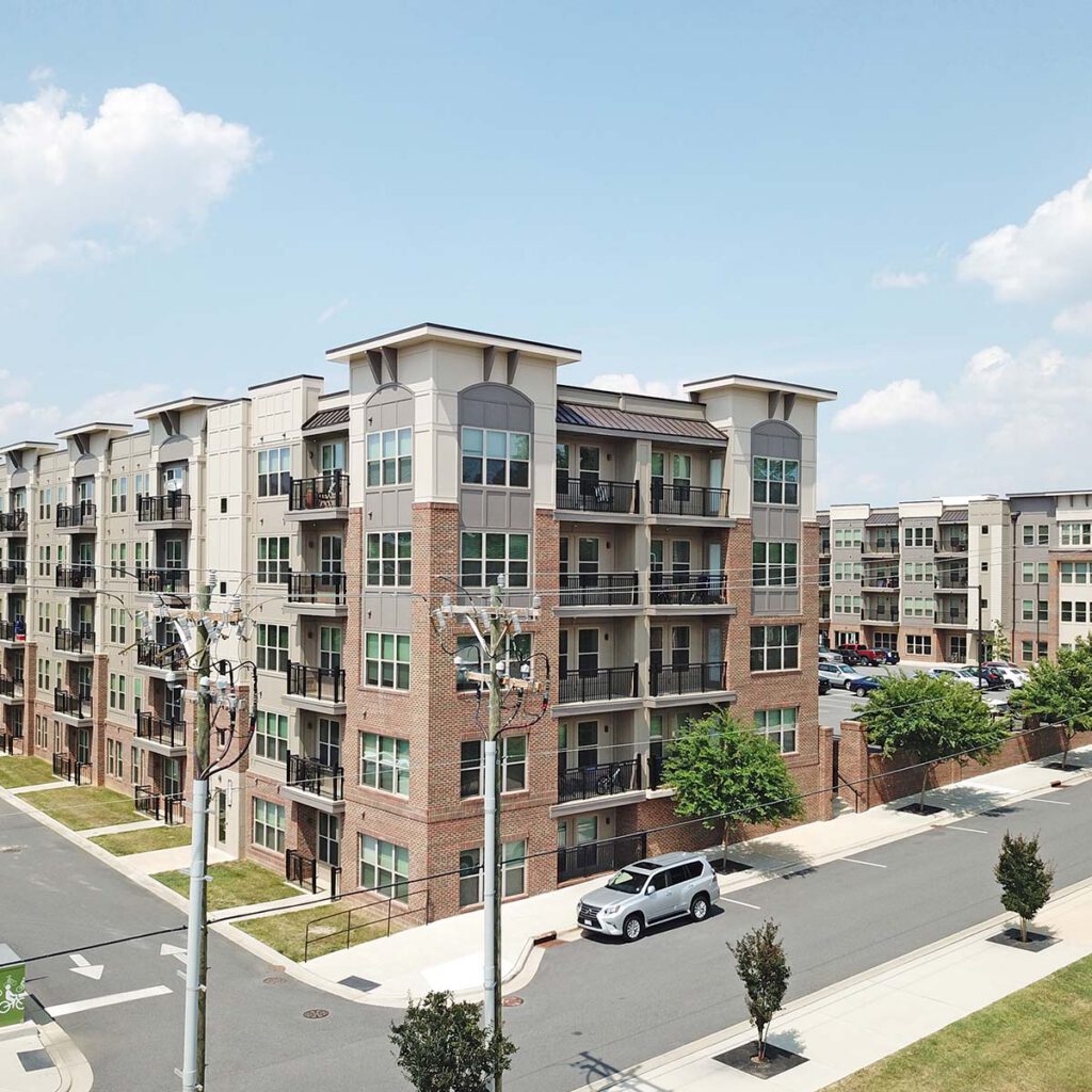 A modern multi-story apartment building with balconies on each floor, a white car parked on the street, and a parking lot with several cars in the background under a clear, blue sky.