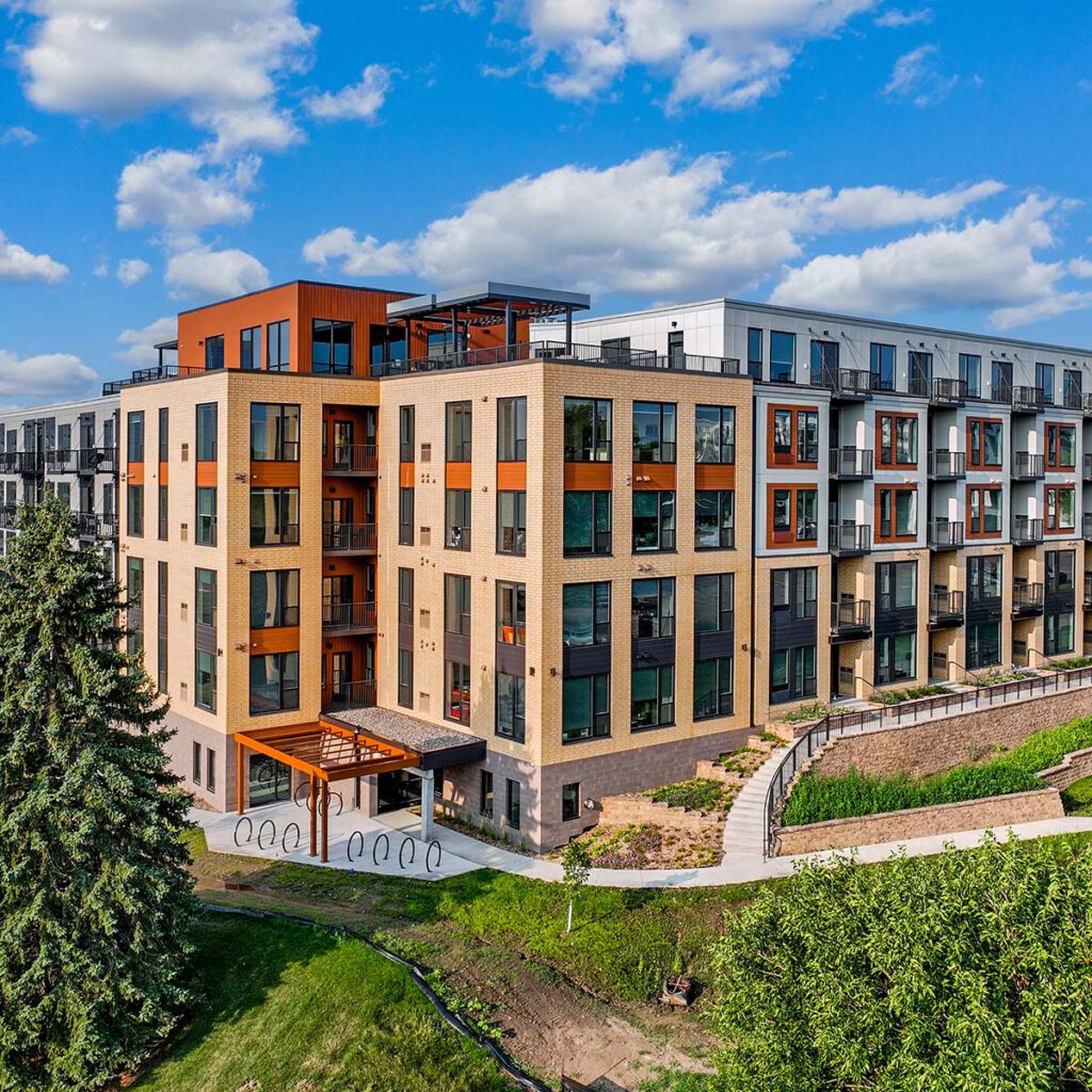 Modern apartment building with large windows, balconies, and a rooftop area, surrounded by green landscaping and a winding sidewalk under a blue sky with scattered clouds.