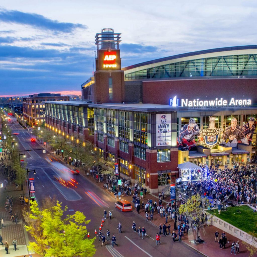 A crowd gathers outside Nationwide Arena in Columbus, Ohio at dusk, with bright lights, large banners, and busy city streets filled with people and cars.