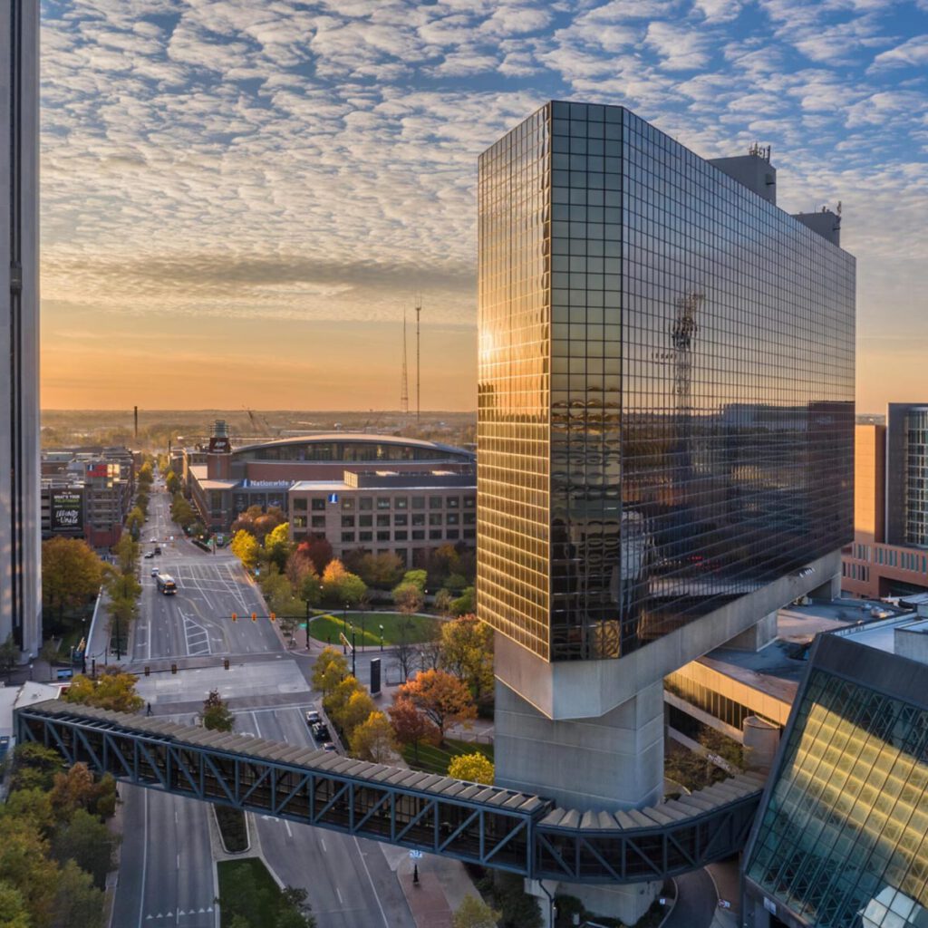 A modern glass skyscraper with a skywalk over a city street at sunrise, reflecting the colorful sky. Nearby are other buildings, a stadium in the background, and autumn trees lining the road.