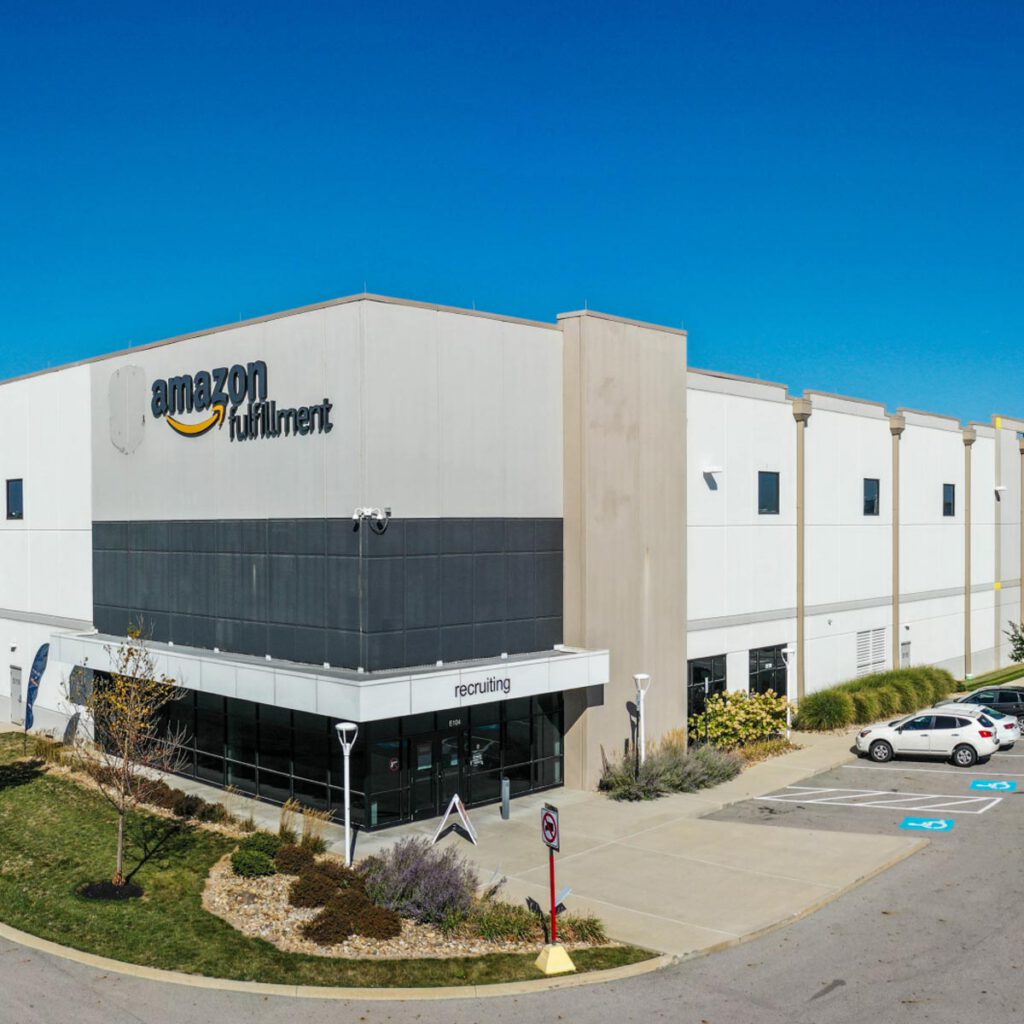 A large Amazon Fulfillment Center building with the Amazon logo on the front, an entrance with a recruiting sign, several parked cars, landscaping, and a clear blue sky overhead.